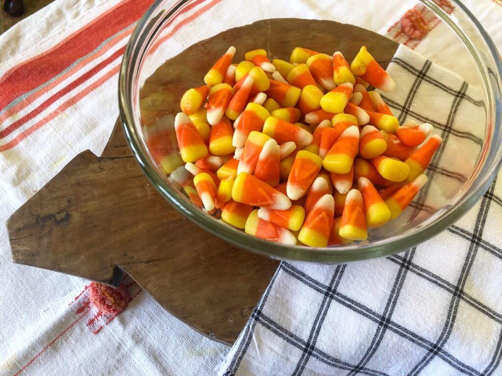 A clear bowl filled with candy corn placed on a wooden cutting board, with a striped and checkered kitchen cloth underneath.