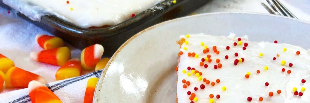 Close-up of a candy corn sheet cake with white frosting and colorful sprinkles, next to candy corn pieces on a checkered tablecloth. A slice of the cake is placed on a plate in the foreground.