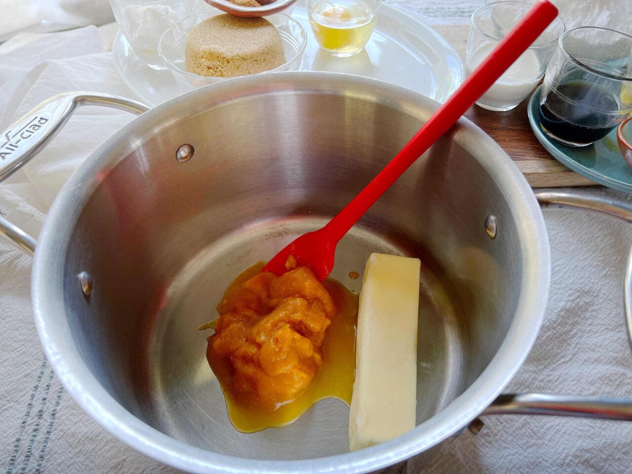 Butter and pumpkin puree in a pot with a red spatula, surrounded by ingredients like brown sugar, vanilla extract, cream, and spices.