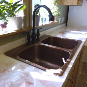 Double bowl copper kitchen sink with bronze faucet in front of a window with plants