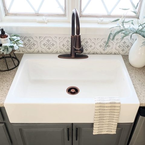 A matte white rectangular Quick-Fit® drop-in farmhouse sink made in fireclay with a bronze faucet, gray cabinets and tan countertop with large window.