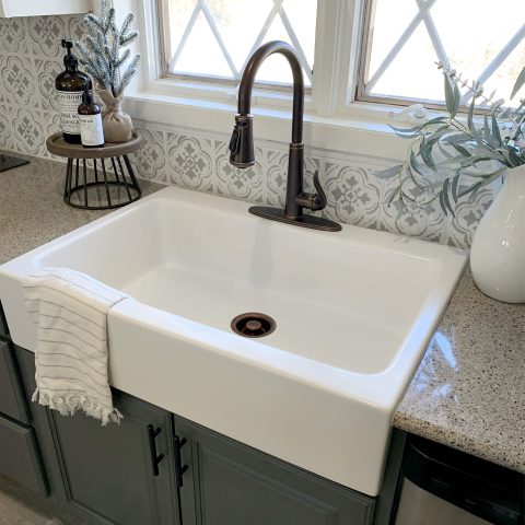 A matte white rectangular Quick-Fit® drop-in farmhouse sink made in fireclay with a bronze faucet, gray cabinets and tan countertop with large window.
