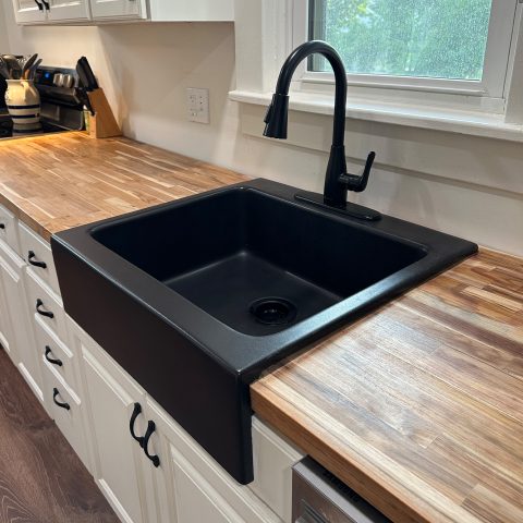 A kitchen with a matte black rectangular Quick-Fit® drop-in farmhouse sink made in fireclay with a black faucet, butcher block countertops and white cabinets.