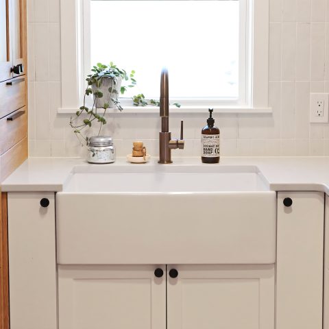 A kitchen with a matte white rectangular undermount farmhouse sink made in fireclay, in front of a window, set in a solid surface white countertop with a modern pull-down faucet and white cabinets.