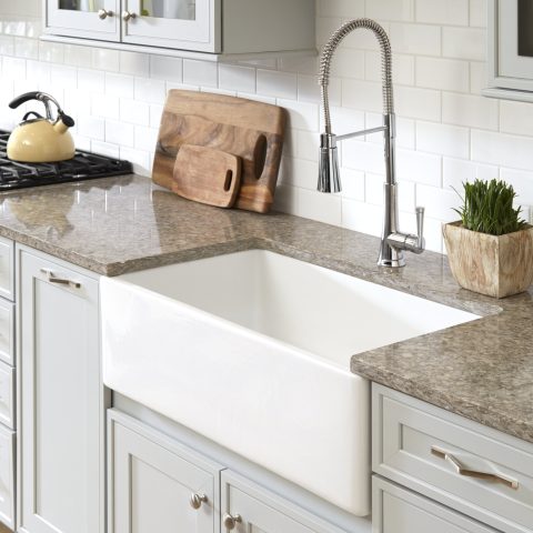 A kitchen with a glossy white rectangular undermount farmhouse sink made in fireclay set in a granite countertop with a chrome pull-down faucet and white tile backsplash.