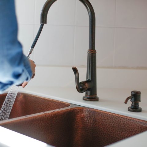 A copper rectangular undermount double basin sink. With a person spraying the sink with the faucet pull down.