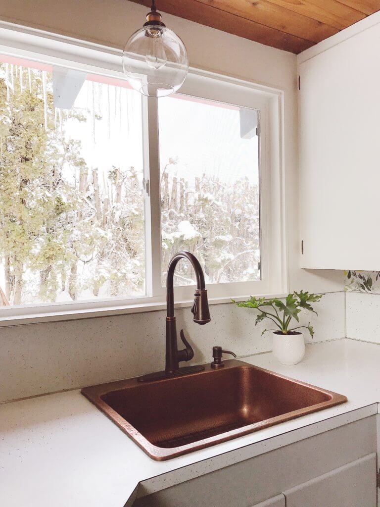 A kitchen sink area with a copper sink and dark faucet, a large window with a snowy view, and a small plant on the white countertop.