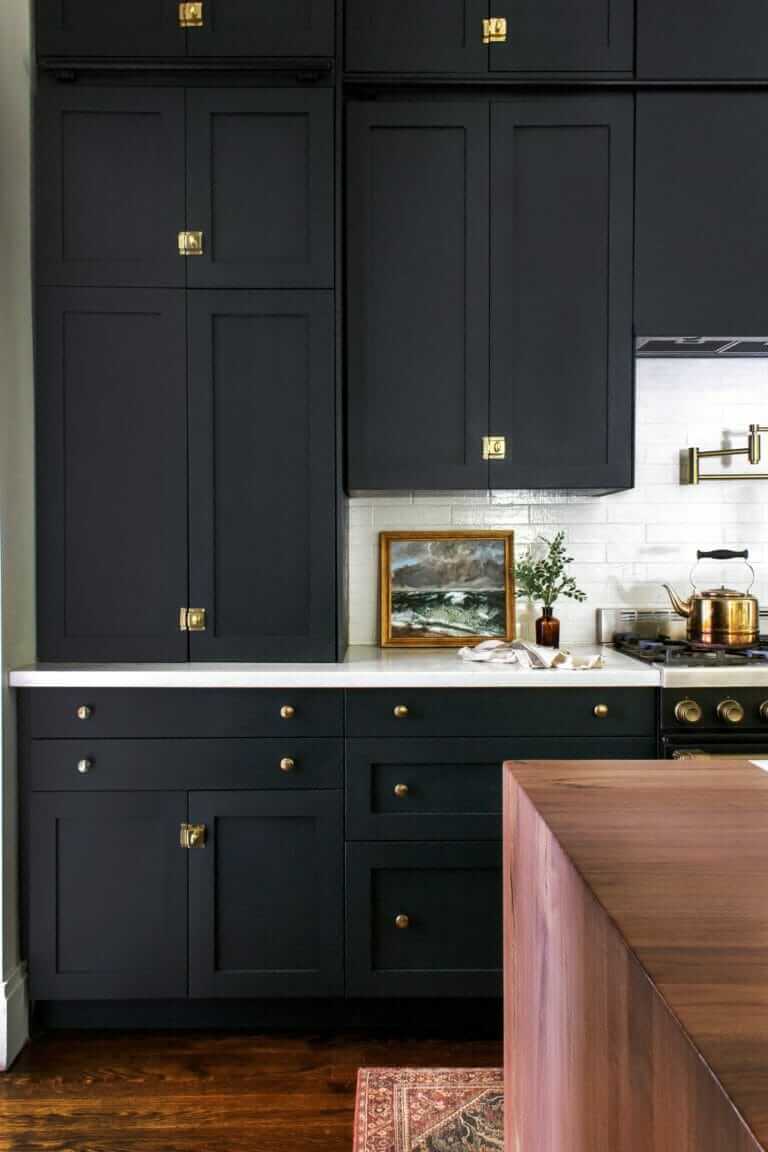 A kitchen with dark cabinetry, brass hardware, a white countertop, and a framed painting placed on the counter near a stove with a brass kettle.