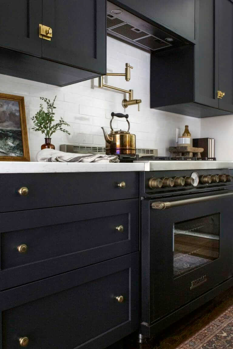 A kitchen stove with brass accents and a brass kettle, set against a white subway tile backsplash, with a pot filler above. The countertop features a small framed painting, a potted plant, and a folded dish towel.