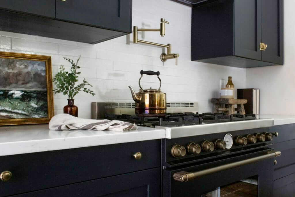 A kitchen stove with brass accents and a brass kettle, set against a white subway tile backsplash, with a pot filler above. The countertop features a small framed painting, a potted plant, and a folded dish towel.