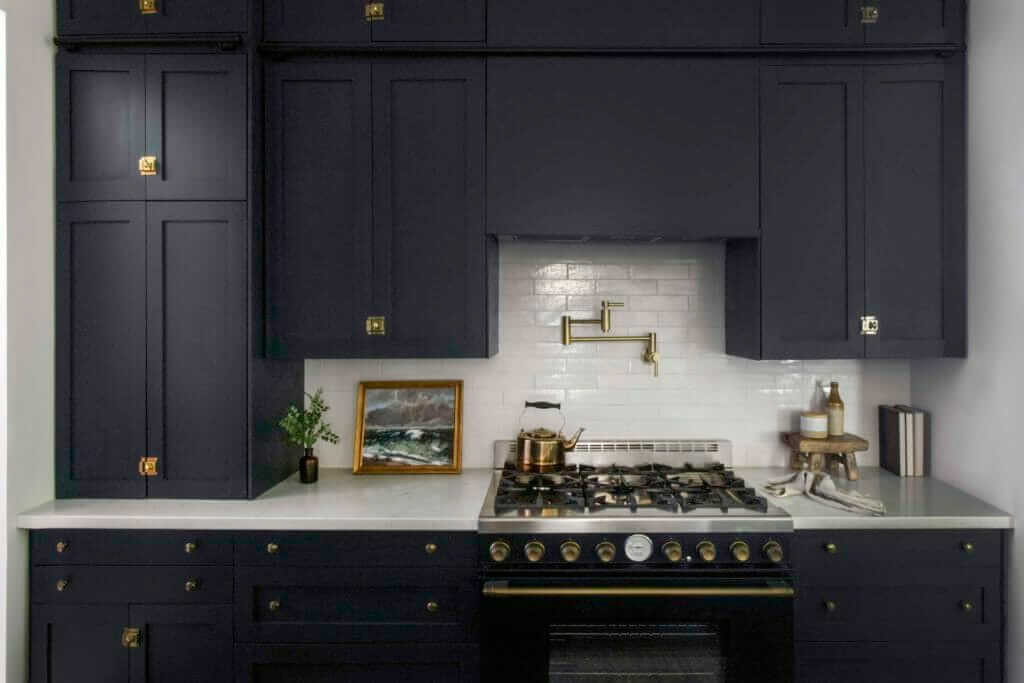 A modern kitchen with dark cabinetry, a white subway tile backsplash, a brass pot filler, and a stove with brass accents. The countertop features a small framed painting, a potted plant, and various kitchen items.