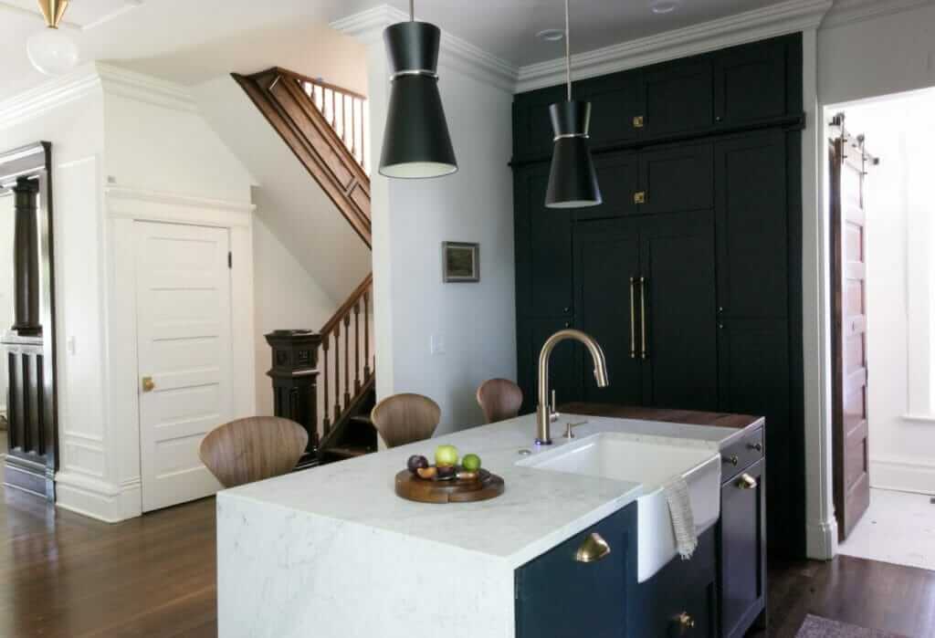 A modern kitchen with a white marble island featuring a farmhouse sink, dark cabinetry, and pendant lights. In the background, there is a staircase with wooden railings and a small wood-burning stove.