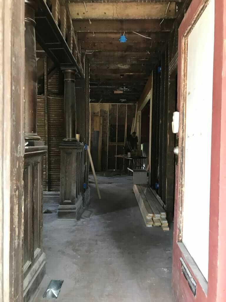 A view into a house under renovation, showing exposed wooden beams and columns, with construction materials like lumber and tools scattered on the floor.