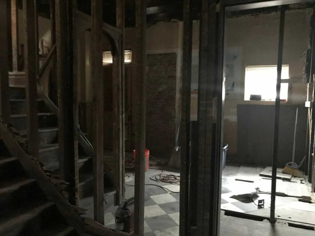 A dark, partially gutted room in a house under renovation, with exposed wall framing, construction debris, and a staircase visible on the left.
