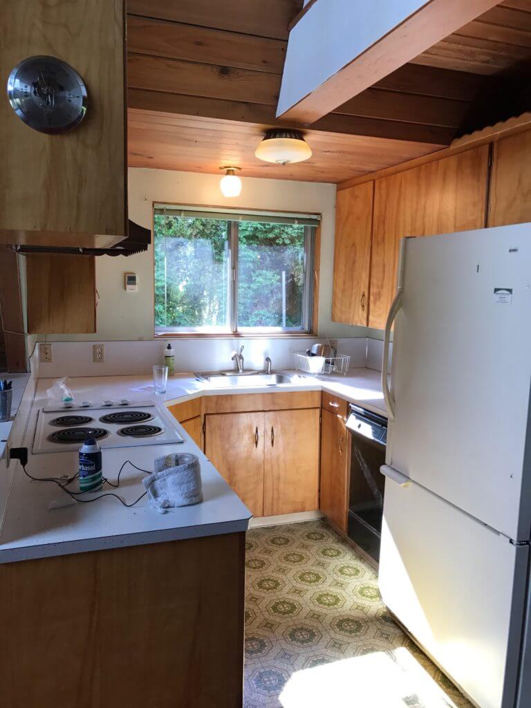A retro kitchen with wooden cabinets, white countertops, an electric stove, and a white refrigerator. The space features a large window, wooden ceiling, and patterned linoleum flooring.